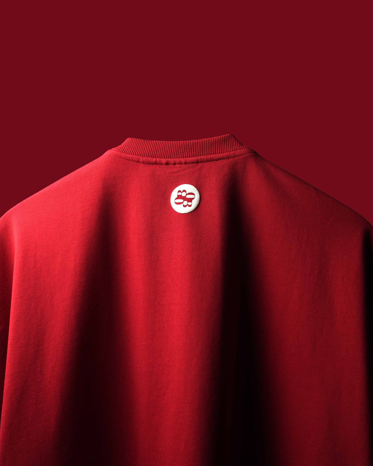 A close-up of a red crewneck T-Shirt showing the back collar with a small circular white and red logo patch, set against a solid dark red background.