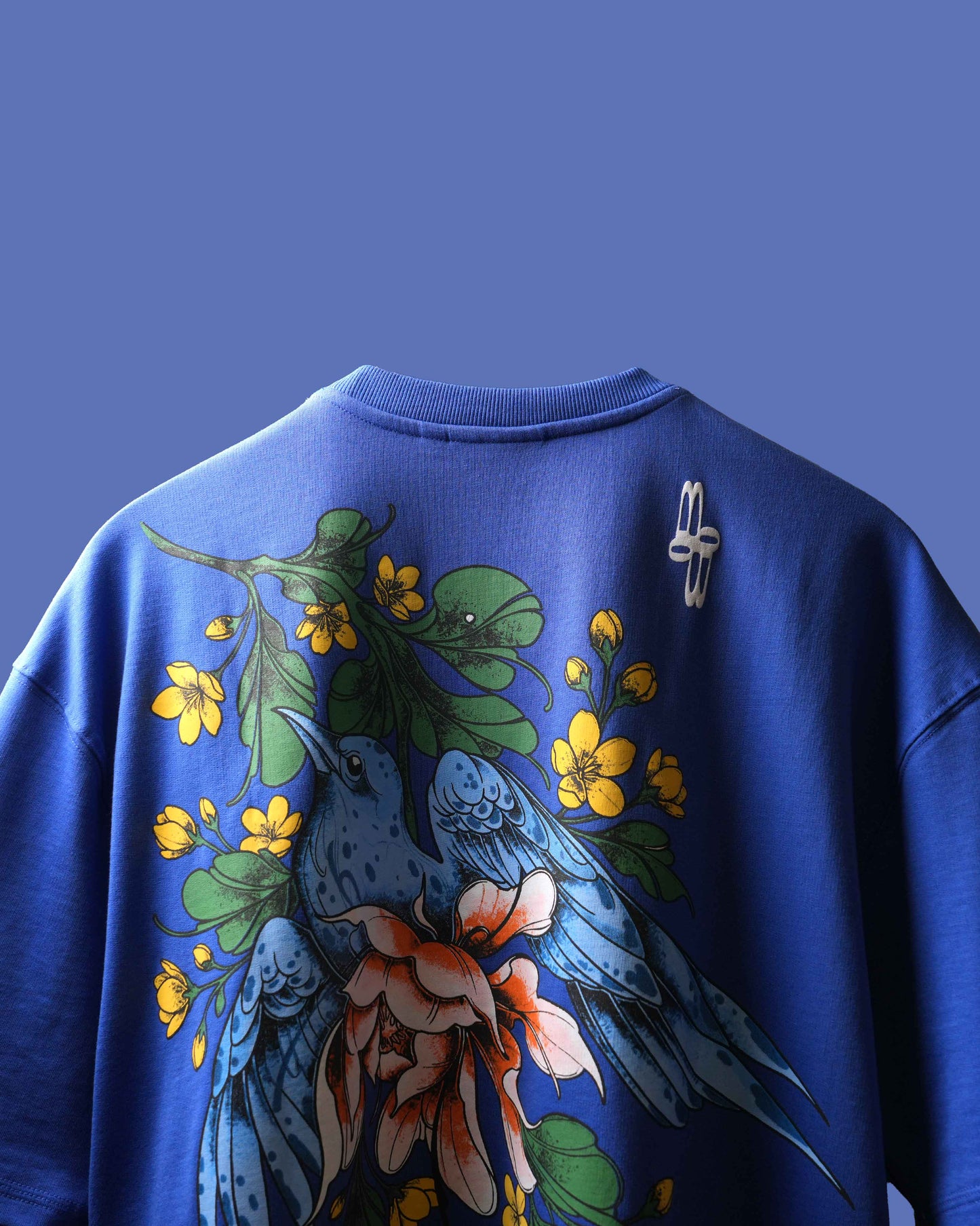 A royal blue crewneck t-shirt featuring a large graphic print on the back with blue birds, yellow flowers, and green leaves, shown against a solid blue background.
