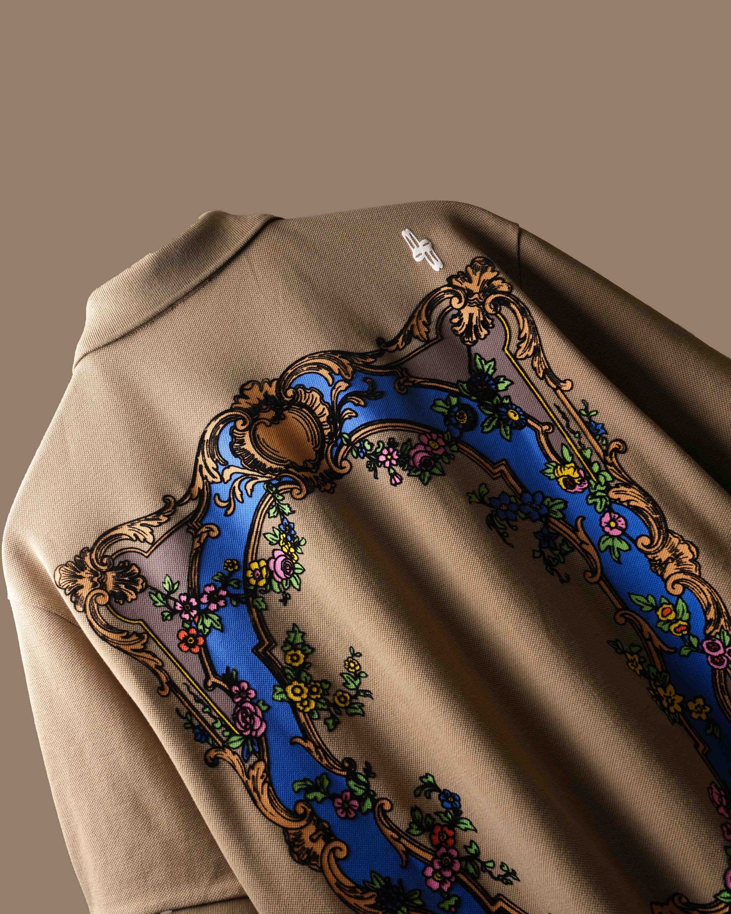 Close-up of the back of a beige polo t-shirt featuring a large ornate baroque-style graphic with a blue arch, gold scrollwork, and floral details.