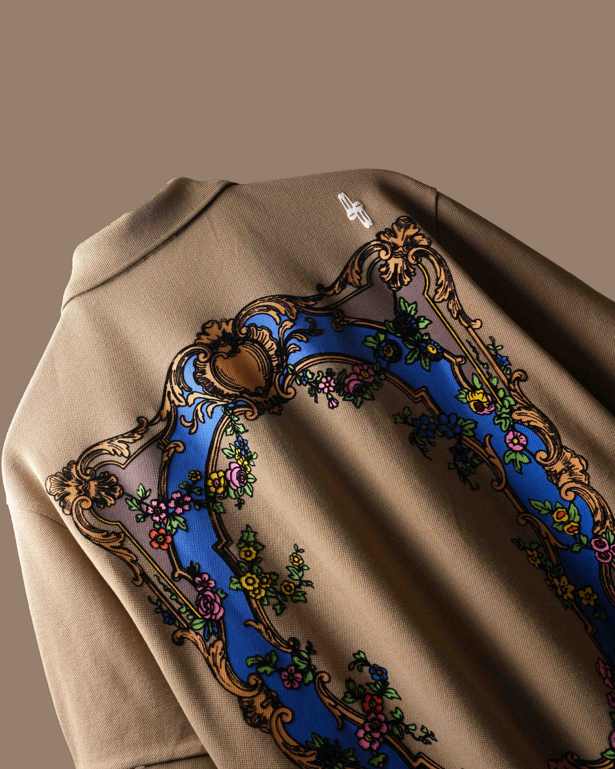 Close-up of the back of a beige polo t-shirt featuring a large ornate baroque-style graphic with a blue arch, gold scrollwork, and floral details.