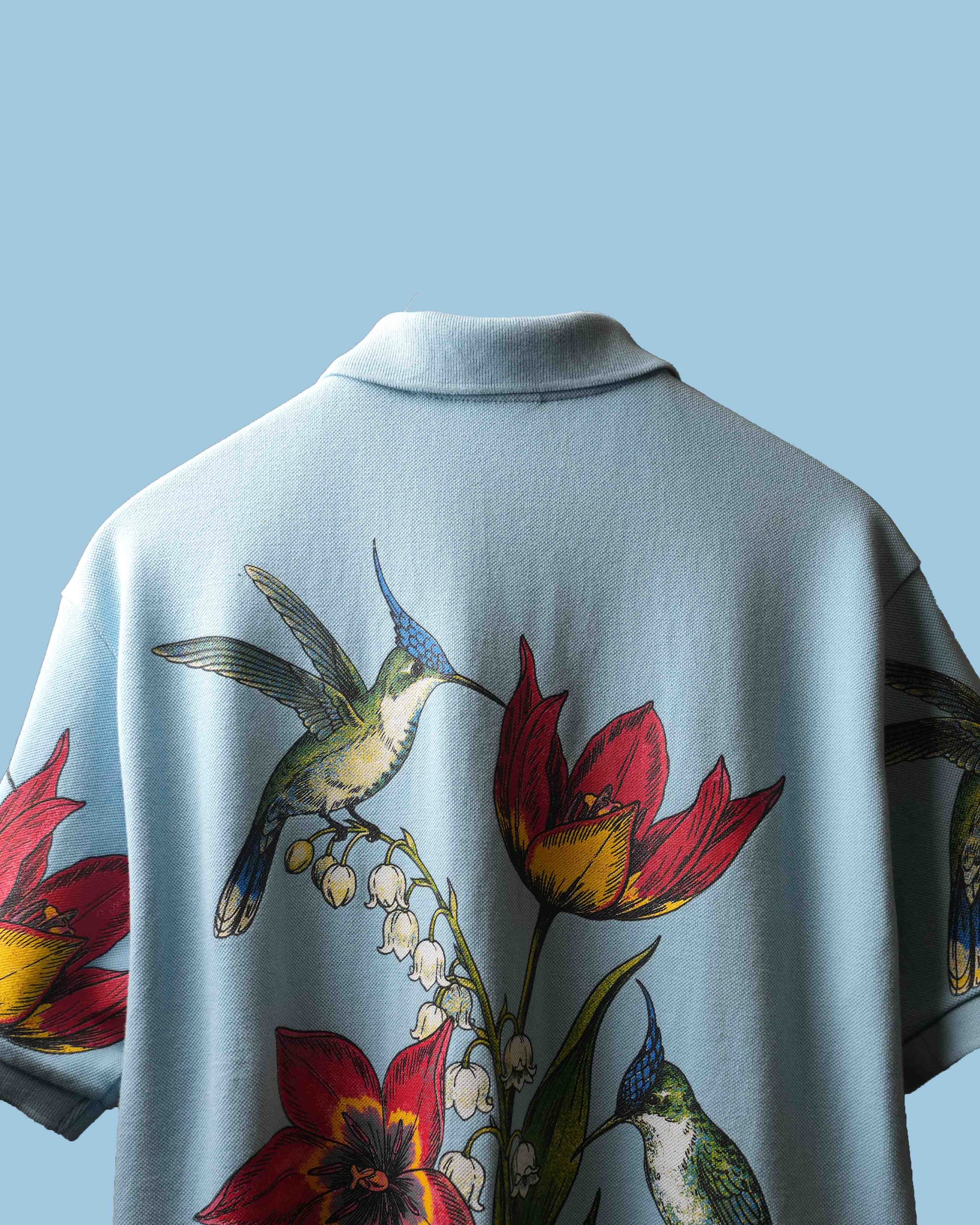 A light blue short-sleeve polo shirt shown from the back, featuring a large colorful print of hummingbirds and red flowers with white lily of the valley blossoms.