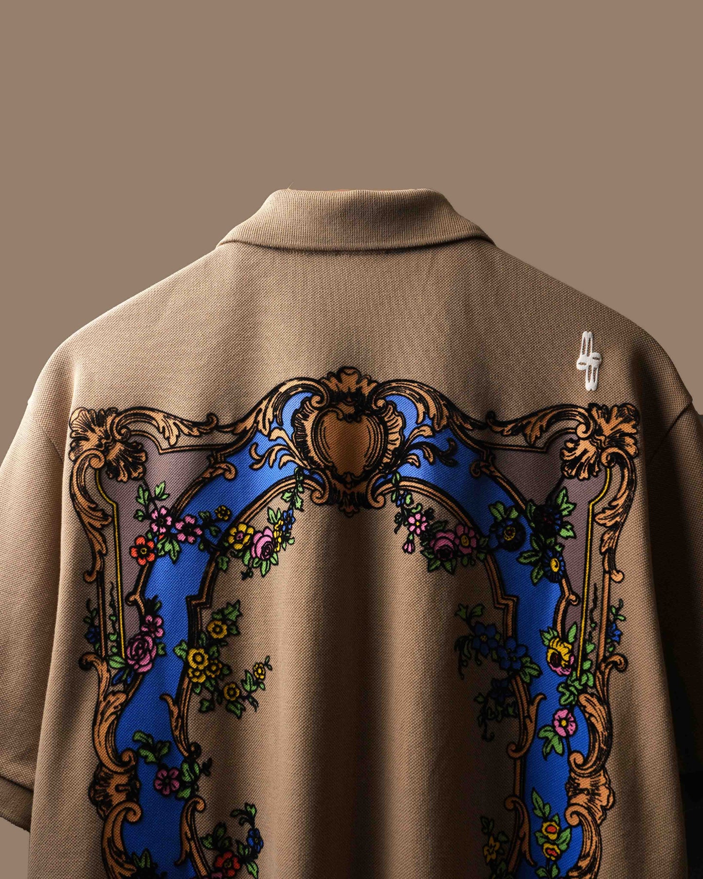 Close-up of the back of a beige polo t-shirt featuring a large ornate baroque-style graphic with a blue arch, gold scrollwork, and floral details.