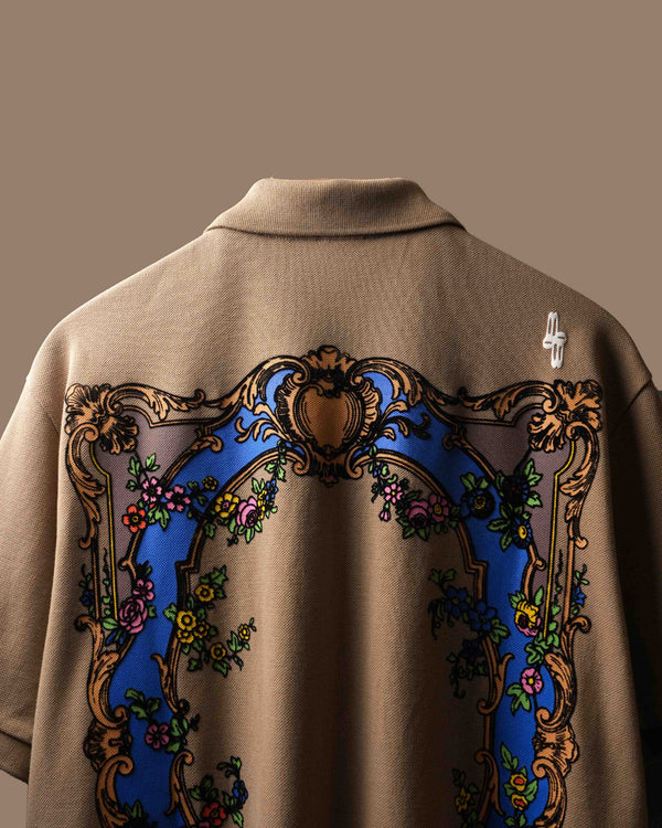 Close-up of the back of a beige polo t-shirt featuring a large ornate baroque-style graphic with a blue arch, gold scrollwork, and floral details.