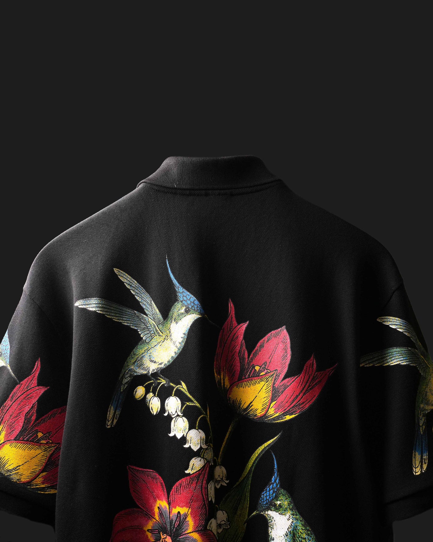 A black short-sleeve Polo featuring a detailed graphic print of a blue and green hummingbird, red flowers, and white lily of the valley on the back, shown against a dark background.