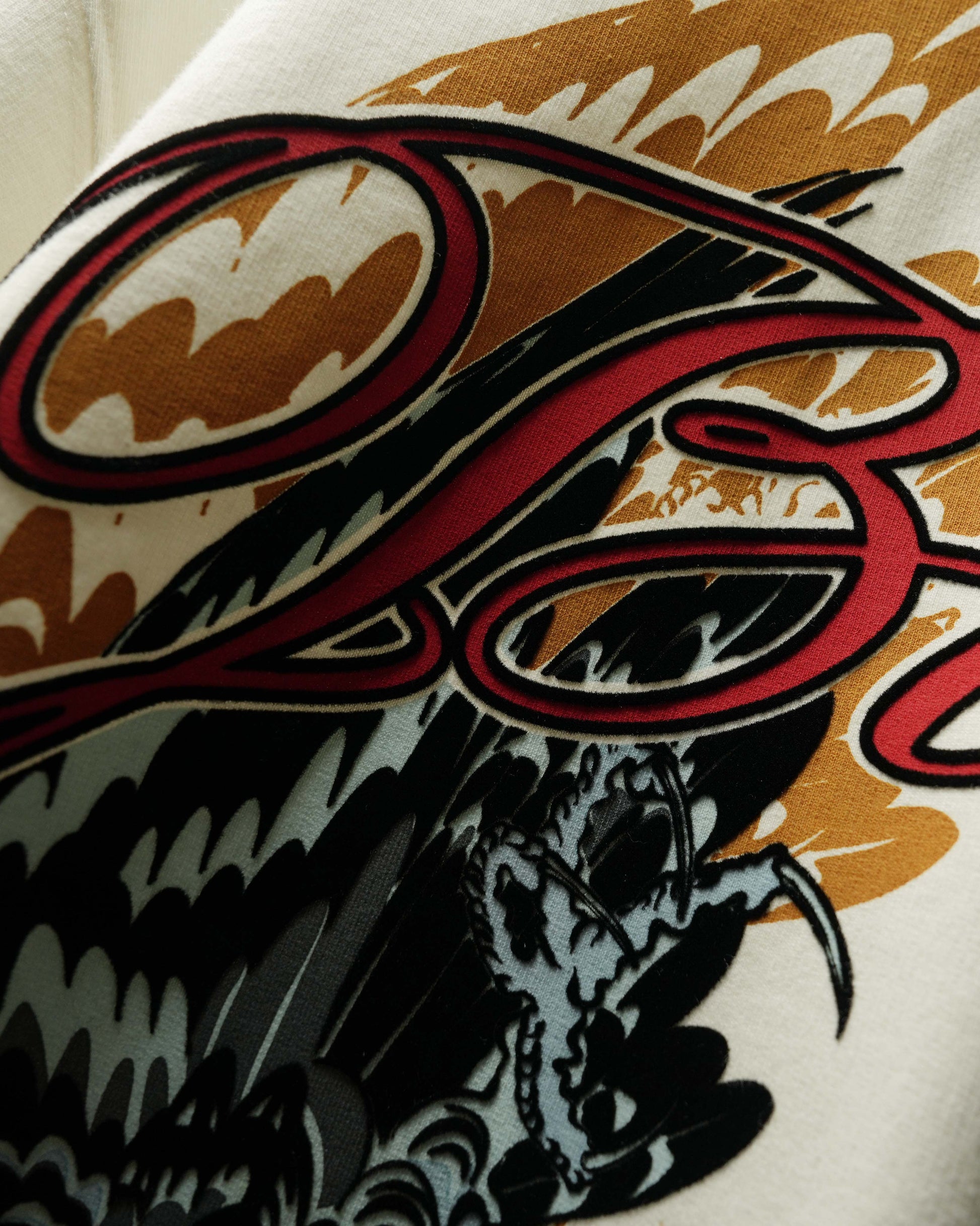 Close-up of a graphic print on a light-colored fabric, featuring a stylized red script logo over black and white illustrated waves and a gray creature, with tan and orange abstract background shapes.