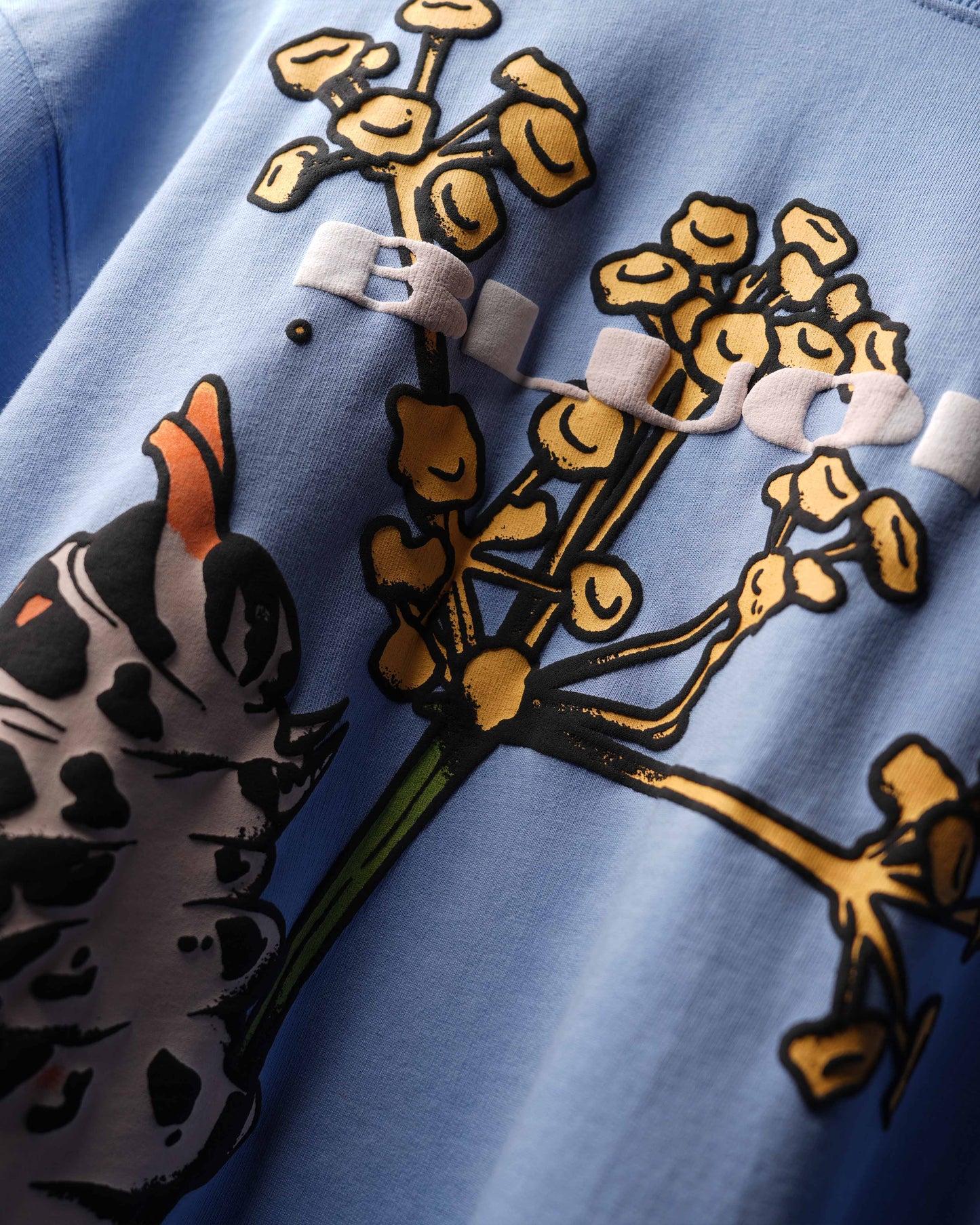 Close-up of a light blue garment featuring a large graphic print of a black-and-white spotted caterpillar and yellow flowers with green stems.