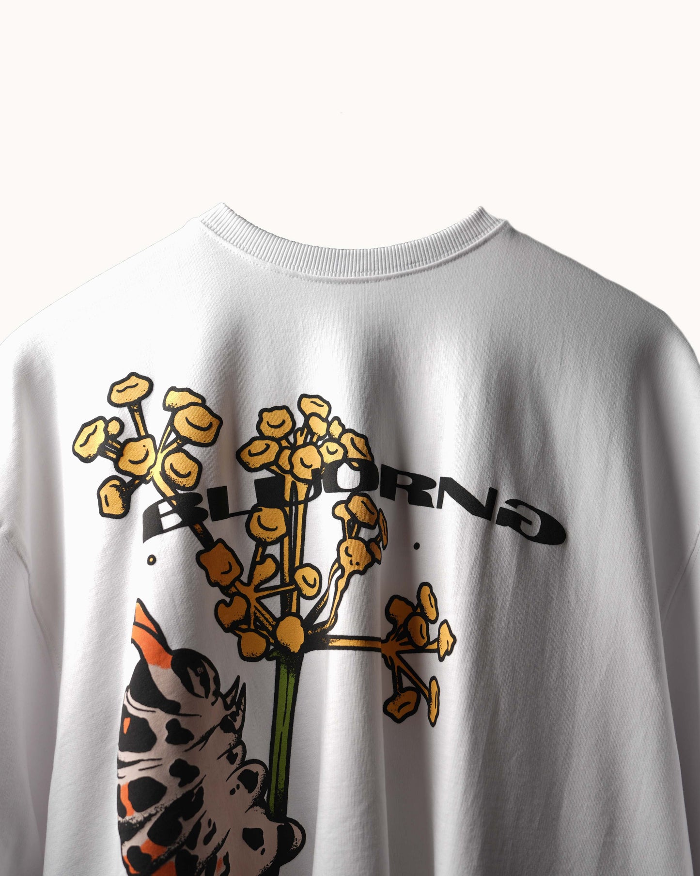A close-up of a white crewneck sweatshirt featuring a graphic print of a yellow-orange flower with the word 'BLOOMING' and a spotted animal face in orange, black, and white.