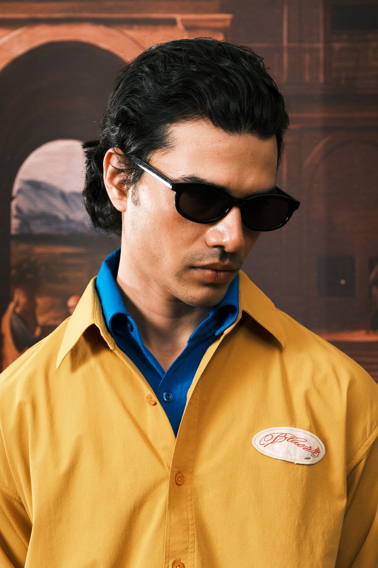 A man wearing black sunglasses, a mustard yellow button-up shirt with a blue collar, and a white oval logo patch on the chest, set against a warm, blurred background.