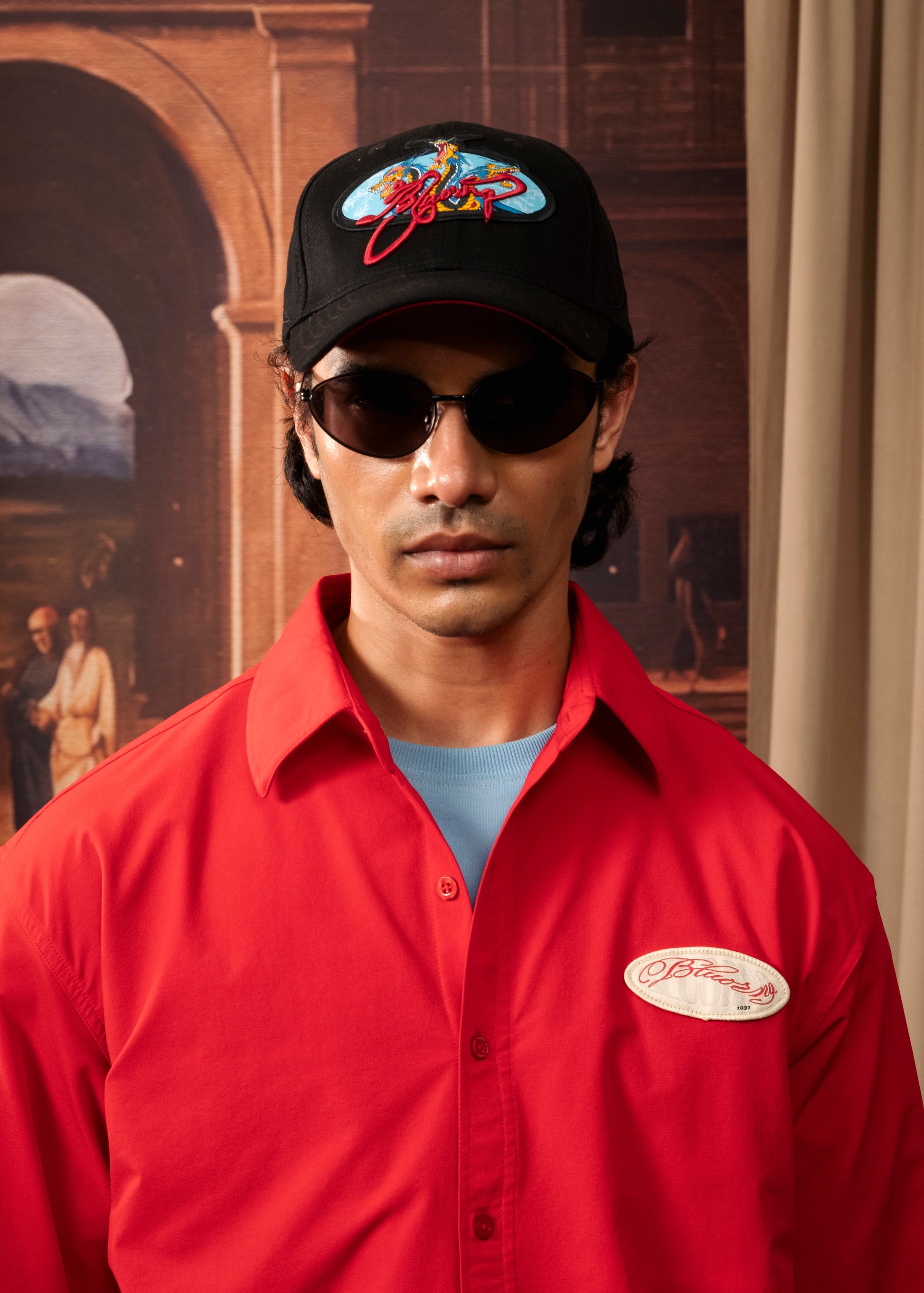 A man wearing a red button-up work shirt with a circular 'Bitter' logo on the left chest, a black baseball cap with a colorful 'Bitter' logo, and dark sunglasses, posed against a warm-toned painted backdrop.