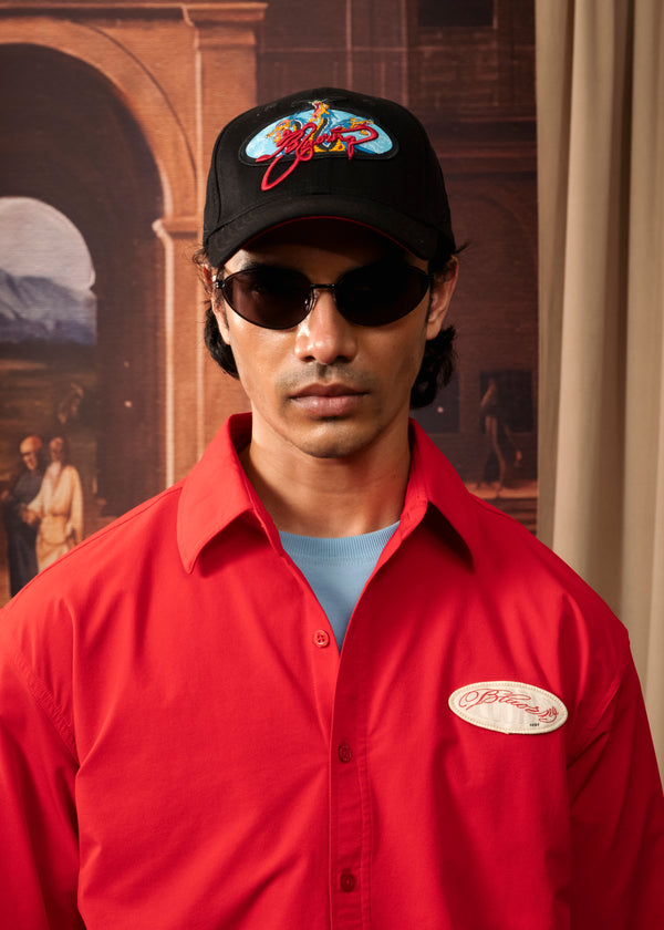 A man wearing a red button-up work shirt with a circular 'Bitter' logo on the left chest, a black baseball cap with a colorful 'Bitter' logo, and dark sunglasses, posed against a warm-toned painted backdrop.
