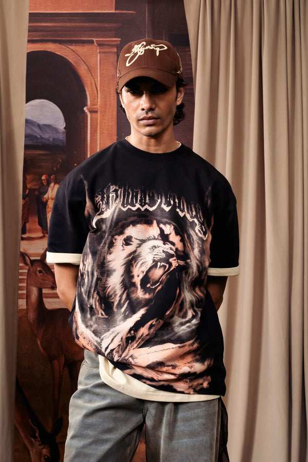 A man wearing a black t-shirt with a large roaring lion graphic, a brown baseball cap, and light gray jeans, standing in front of beige curtains.