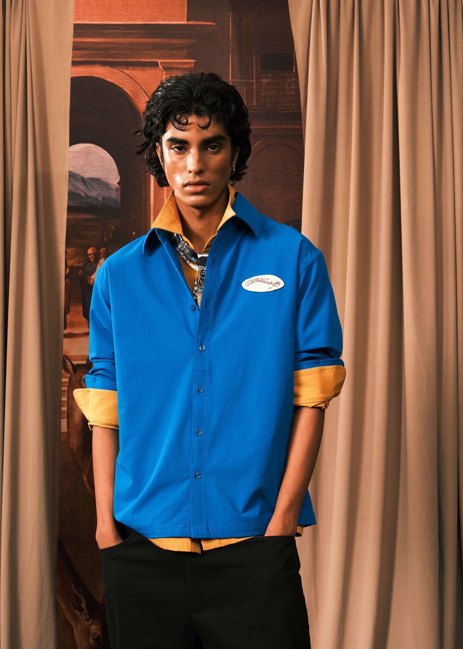 A man wearing a bright blue button-up shirt with rolled-up sleeves showing a tan undershirt, paired with black trousers, standing in front of beige curtains.