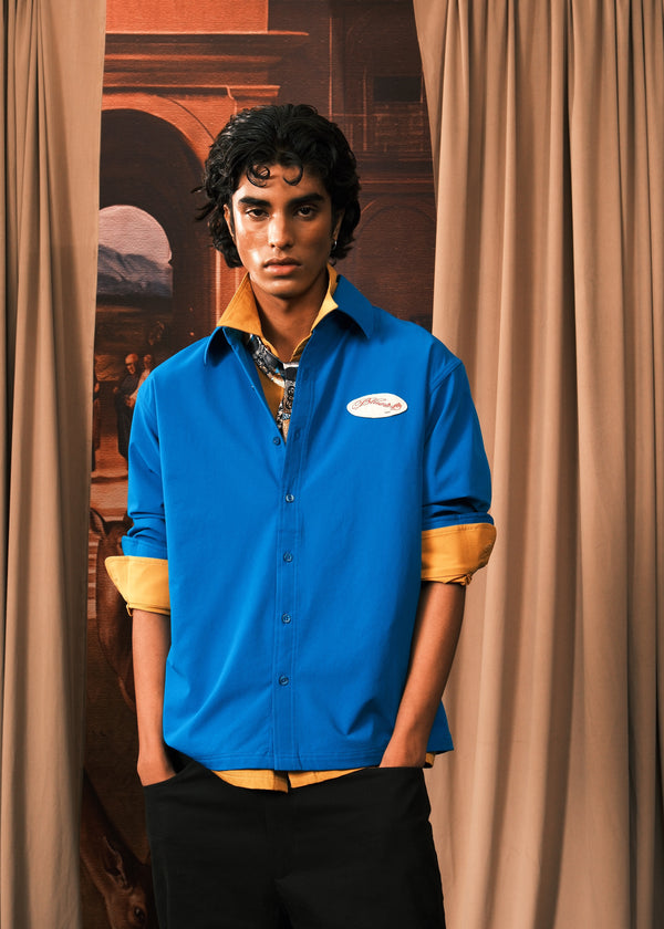 A man wearing a bright blue button-up shirt with rolled-up sleeves showing a tan undershirt, paired with black trousers, standing in front of beige curtains.