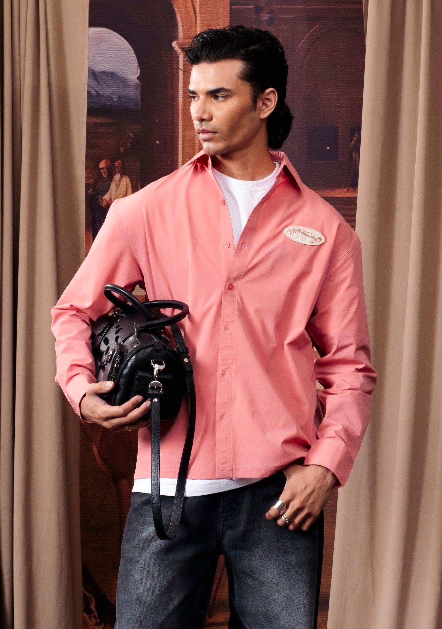 A man stands in front of beige curtains wearing a pink button-up shirt over a white undershirt, dark wide-leg jeans, and holding a black backpack.
