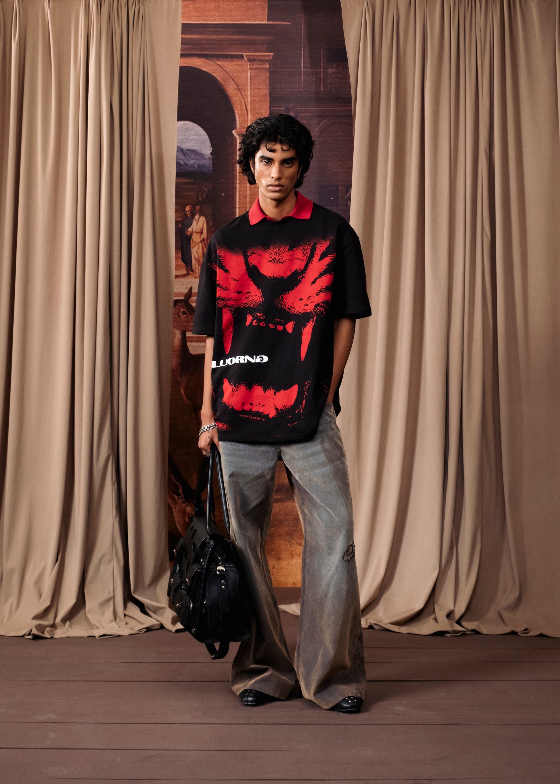 A model stands in a studio setting wearing a black short-sleeve polo shirt with a large red graphic print and the brand name 'LOUNING' on the front, paired with light gray wide-leg jeans and holding a black duffel bag.