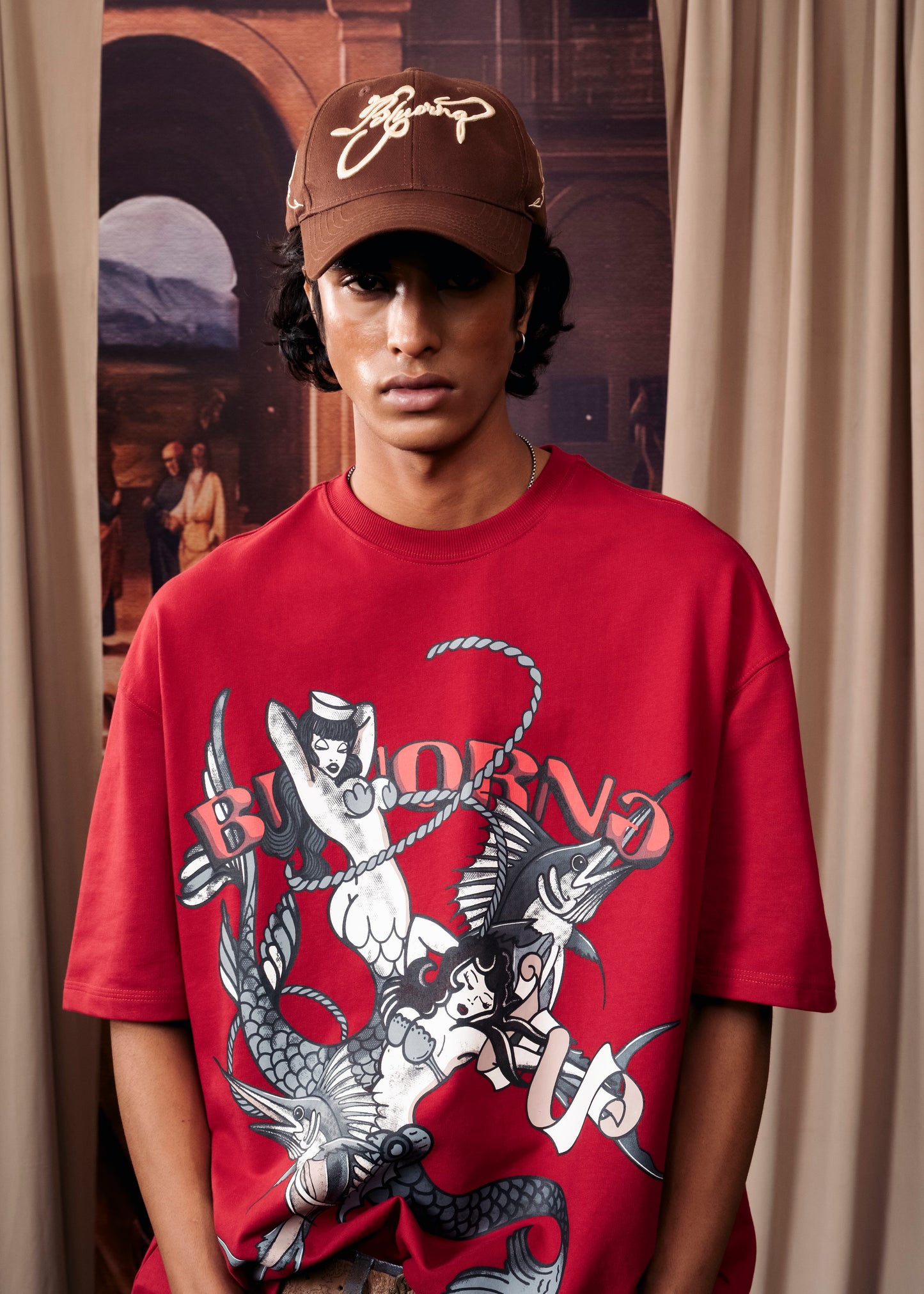 A model wearing a red graphic t-shirt and a brown cap with a white logo, standing in front of a beige curtain and a large art print.