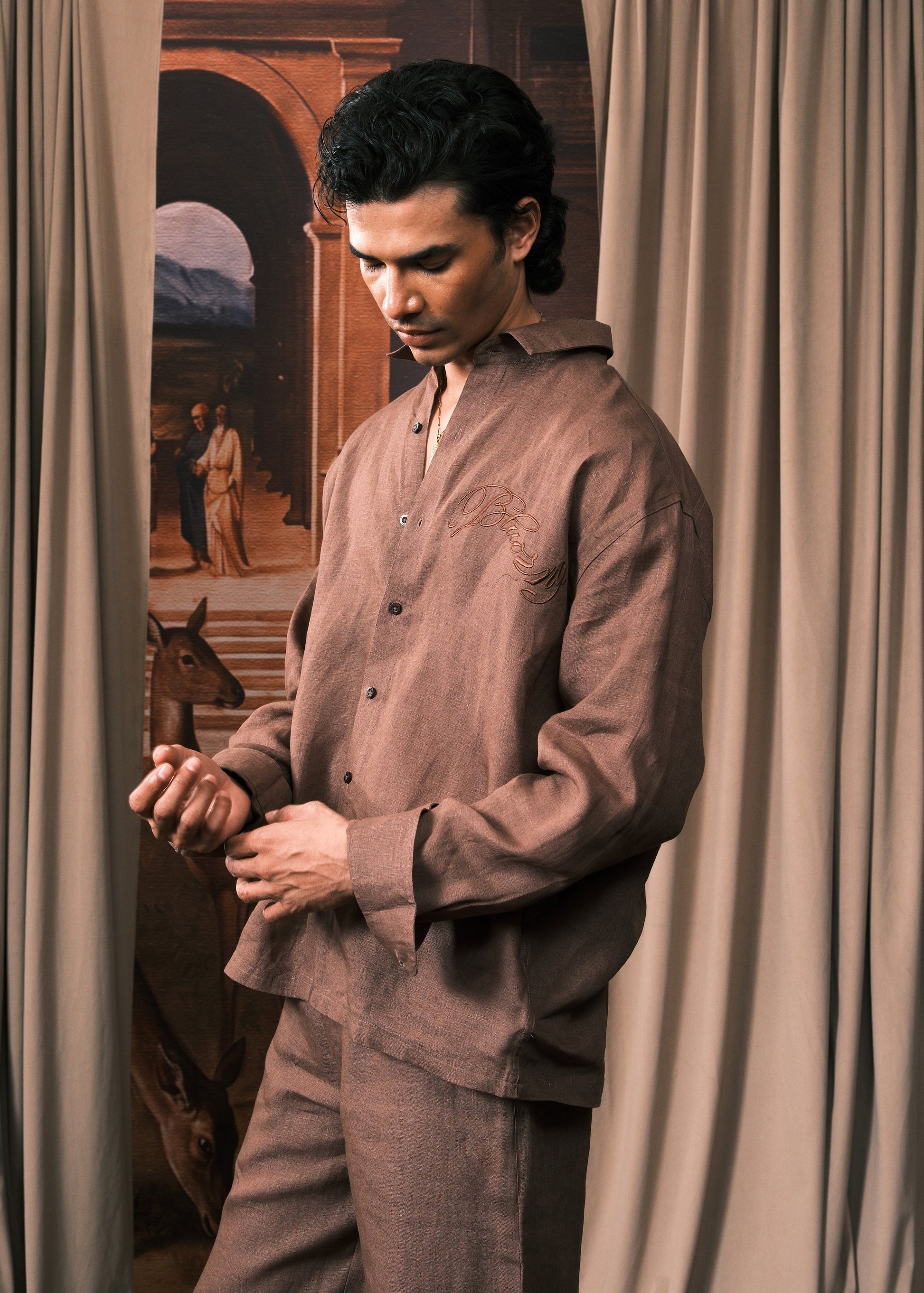 A man wearing a matching brown linen pajama set consisting of a long-sleeve button-up shirt and trousers, standing in front of beige curtains with a vintage painting in the background.