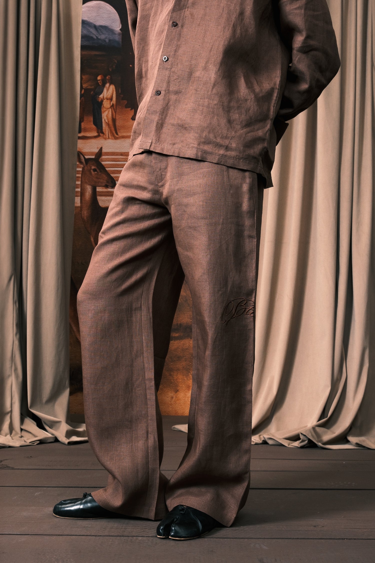 A person wearing a matching brown linen pajama set with a button-up shirt and wide-leg pants, paired with black loafers, standing in front of draped curtains.