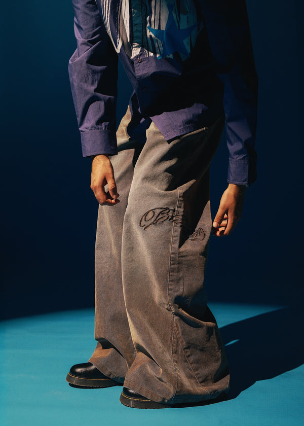 A person wearing very wide-leg brown corduroy Denim with a script logo on the left thigh, styled with a purple shirt and a patterned blue and white top, standing on a blue studio floor.