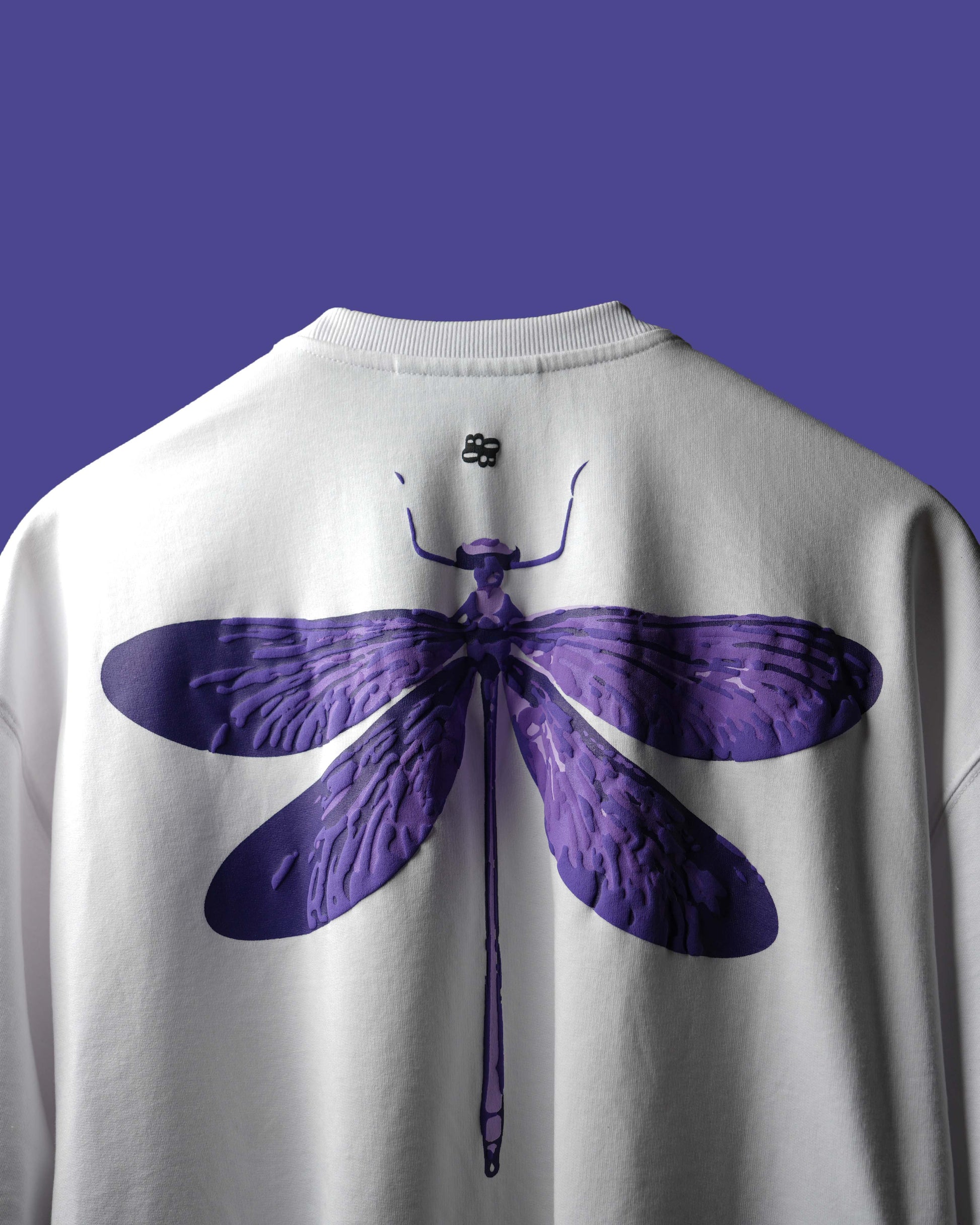 A close-up of the back of a white crewneck sweatshirt featuring a large purple dragonfly graphic print, set against a solid purple background.