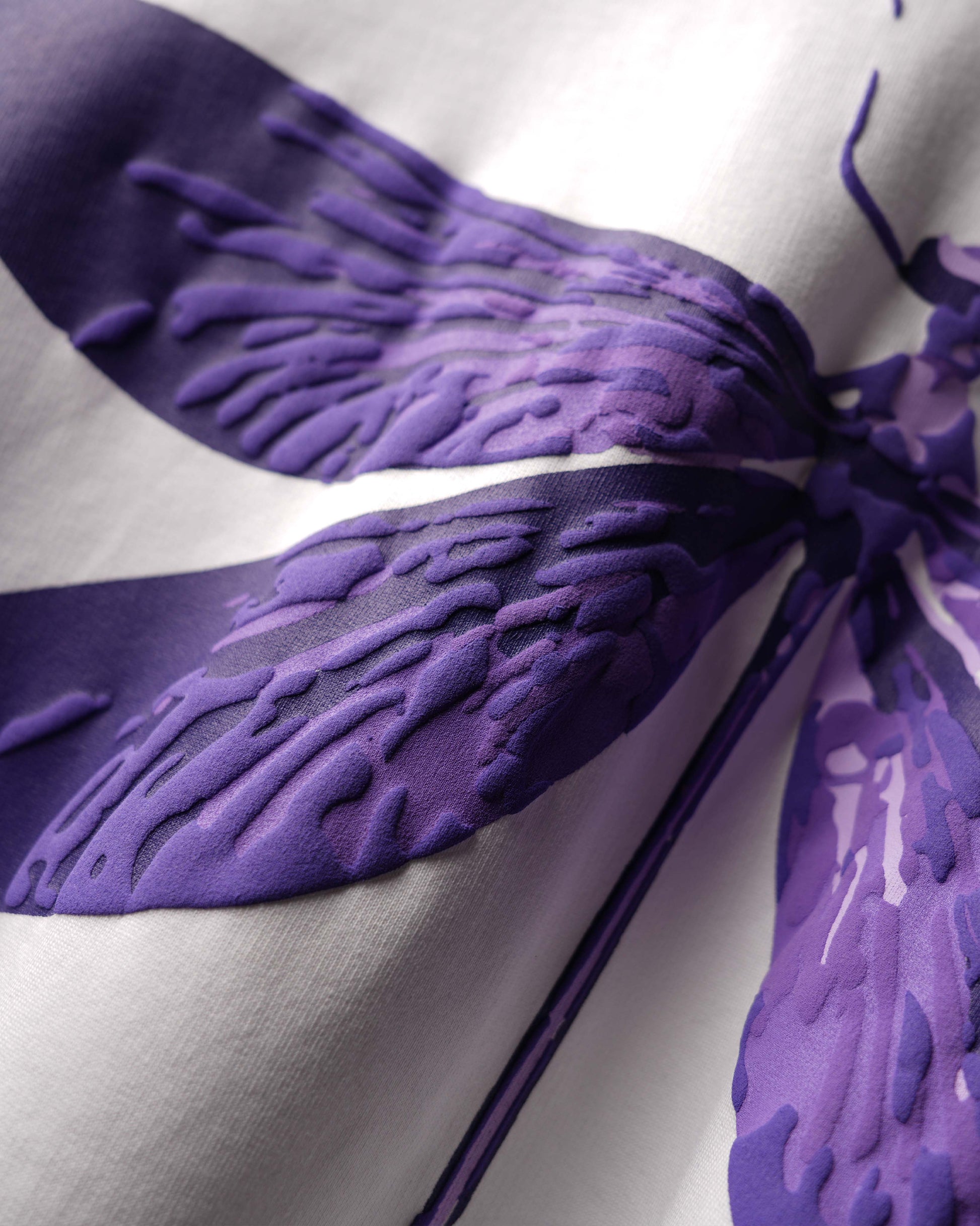 A close-up of a white fabric garment featuring a large purple abstract floral or wing-like pattern with textured, raised details.