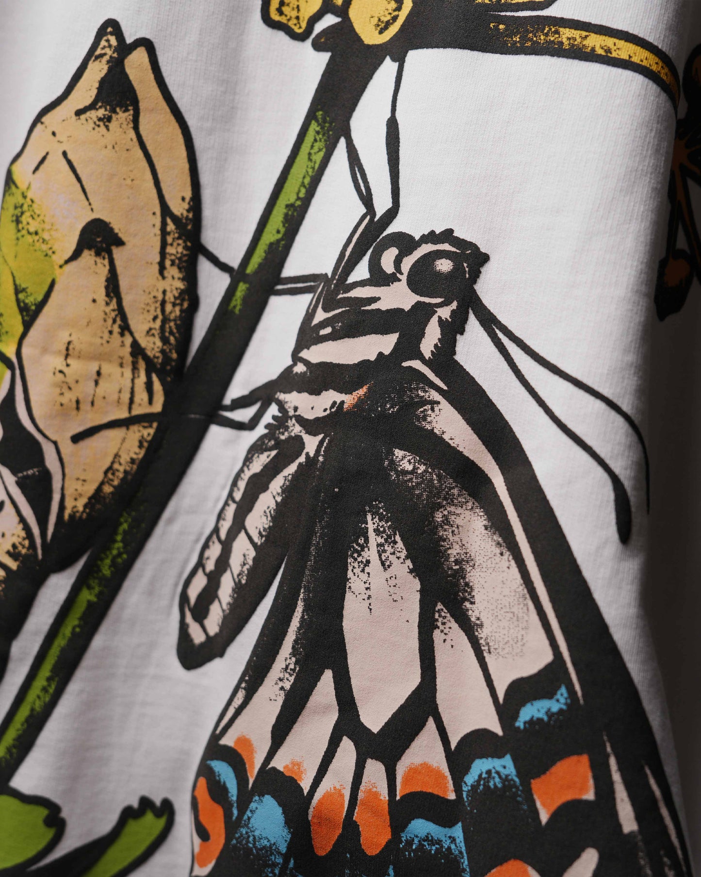 Close-up of a white garment featuring a graphic print of a butterfly on a leaf with black, beige, green, orange, and blue details.
