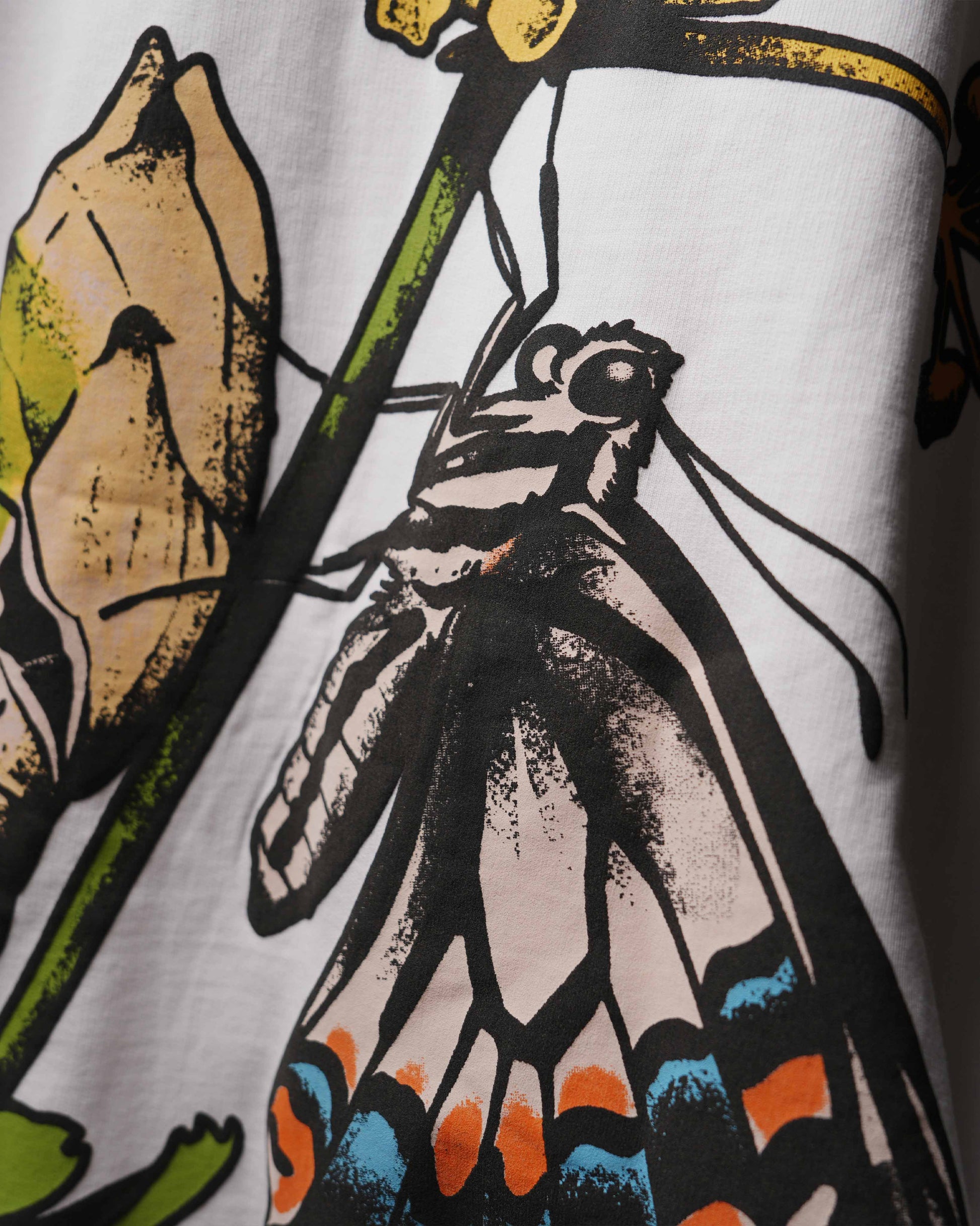Close-up of a white garment featuring a graphic print of a butterfly on a leaf with black, beige, green, orange, and blue details.