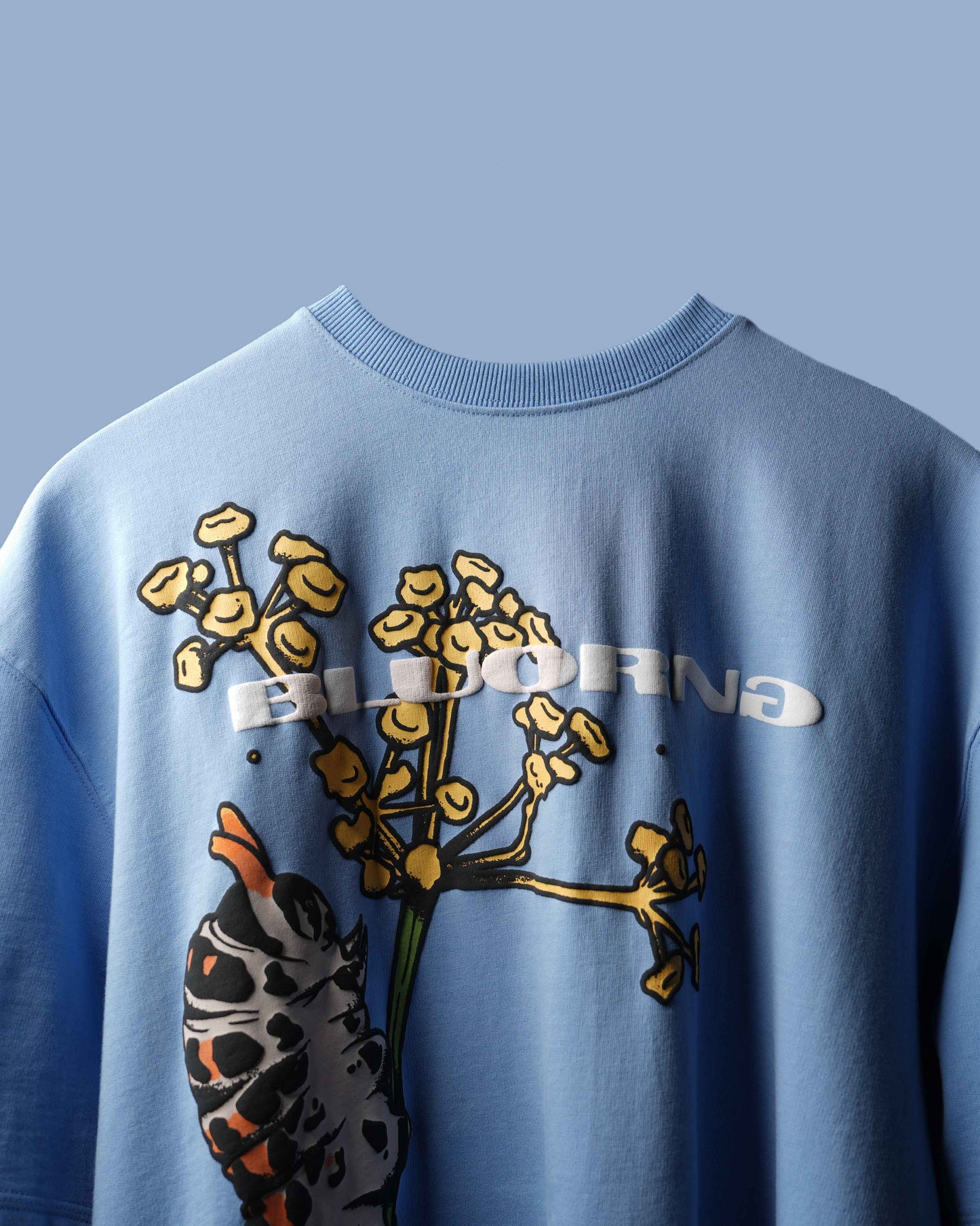 A light blue crewneck sweatshirt featuring a graphic print of a hand holding yellow wildflowers, with the word 'ELDORNA' printed in white across the chest.
