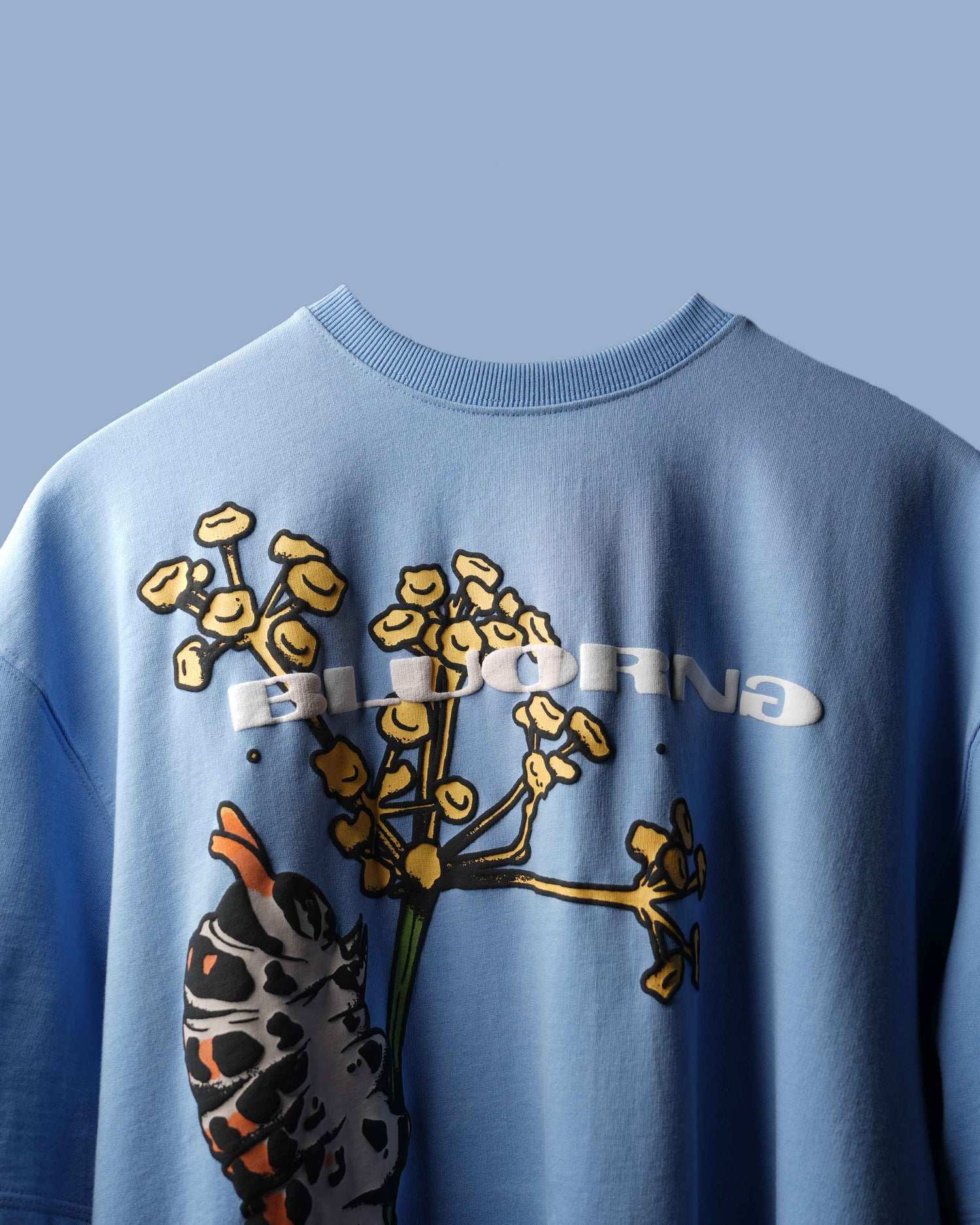 A light blue crewneck sweatshirt featuring a graphic print of a hand holding yellow wildflowers, with the word 'ELDORNA' printed in white across the chest.