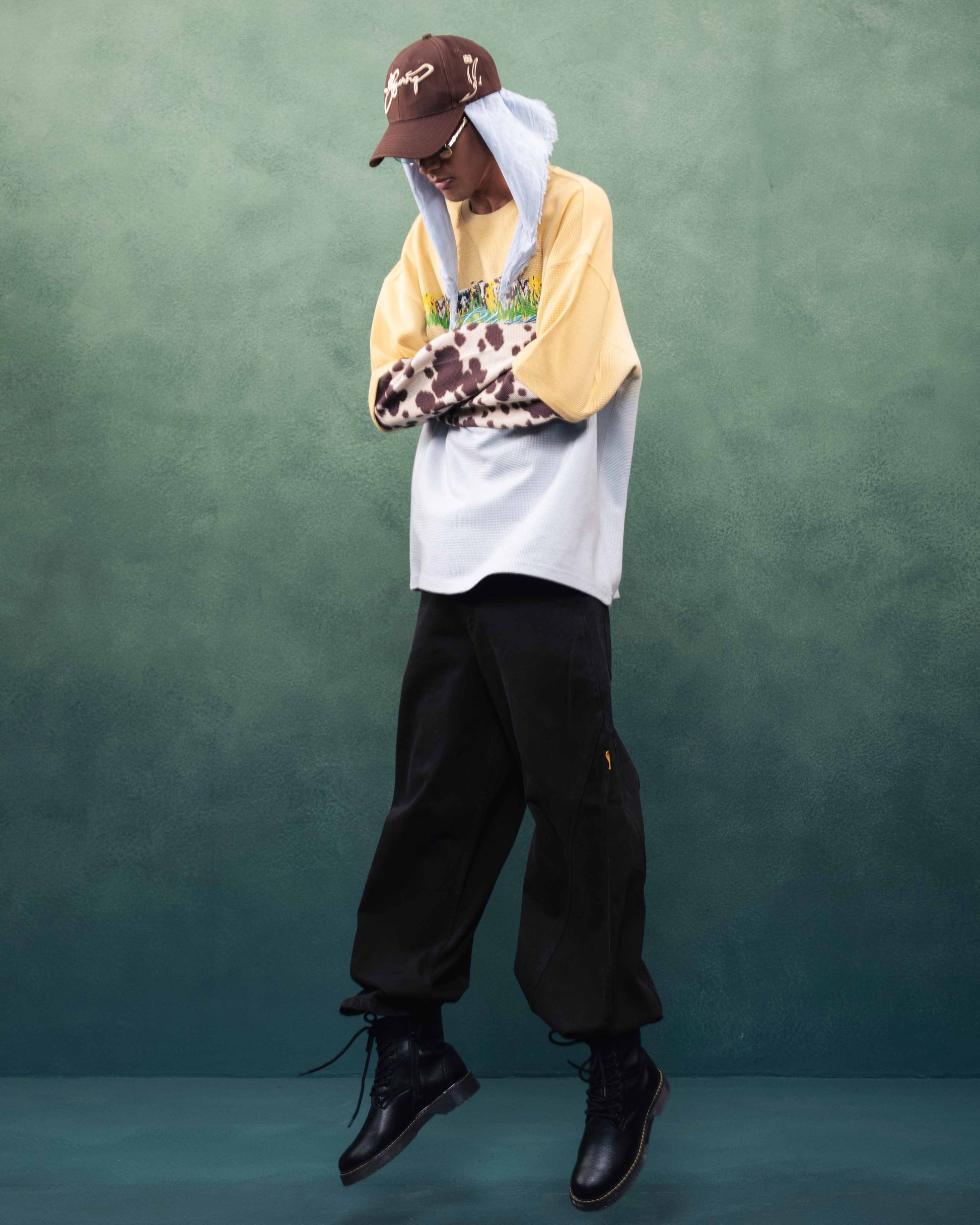 Person wearing a layered outfit with a cap, yellow shirt, white hoodie, black pants, and black shoes against a green background.