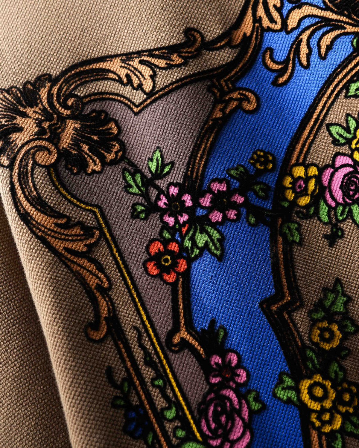 Close-up of the back of a beige polo t-shirt featuring a large ornate baroque-style graphic with a blue arch, gold scrollwork, and floral details.