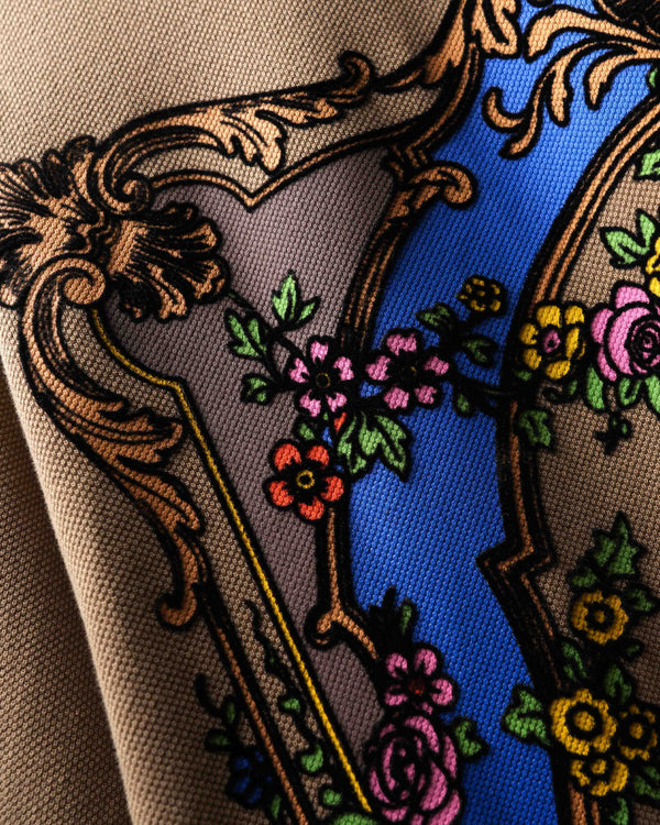 Close-up of the back of a beige polo t-shirt featuring a large ornate baroque-style graphic with a blue arch, gold scrollwork, and floral details.