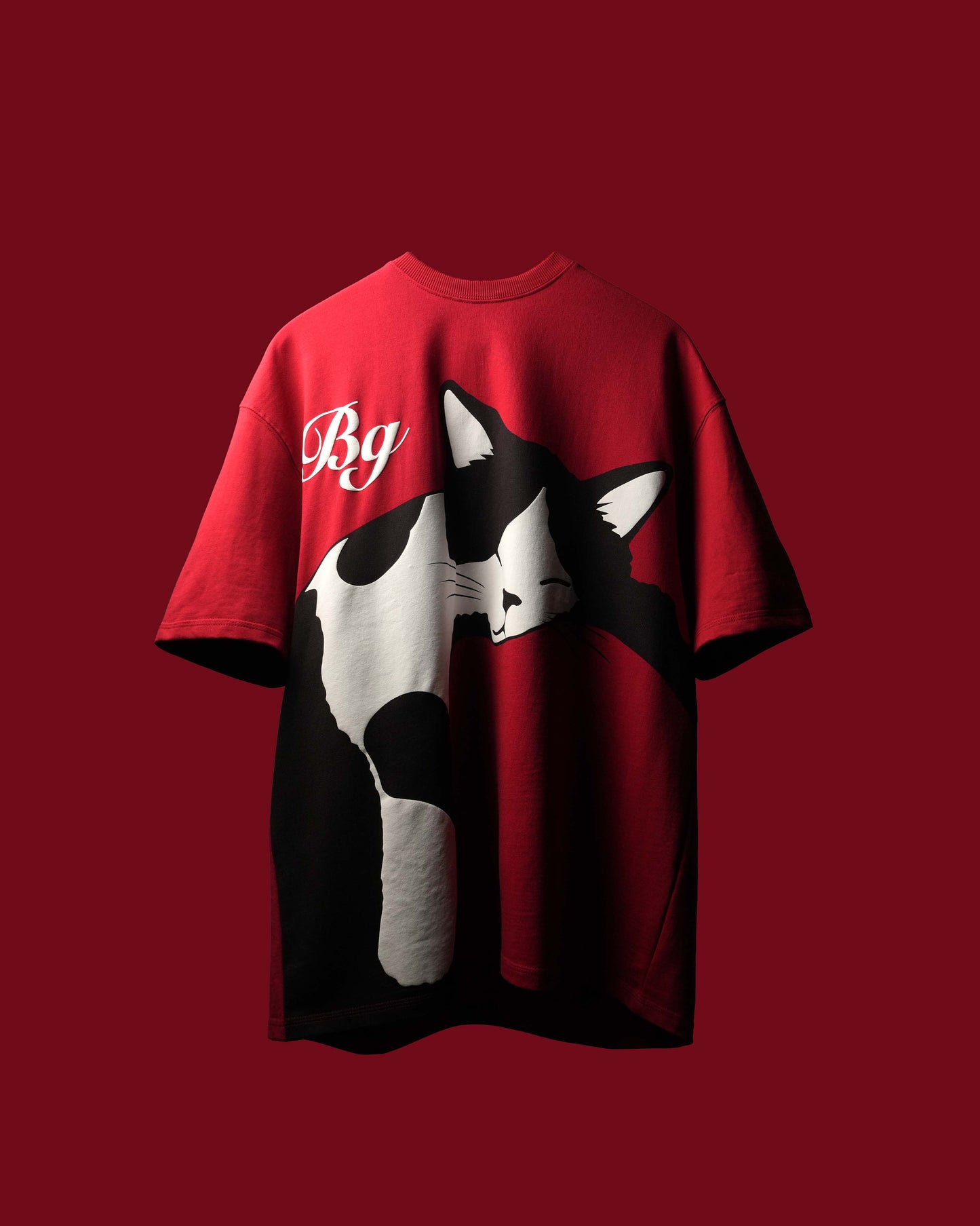 A red short-sleeve t-shirt featuring a large black and white cat graphic on the front and a white cursive 'Bg' logo on the upper left chest, displayed against a solid dark red background.