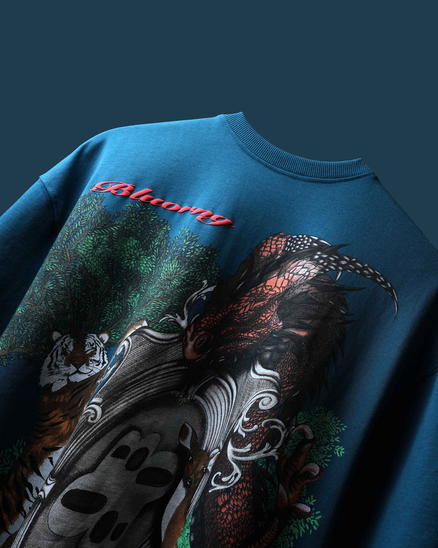 A close-up of a blue crewneck t-shirt featuring a detailed graphic print with a tiger, a red dragon, and green foliage, accented with red script text.
