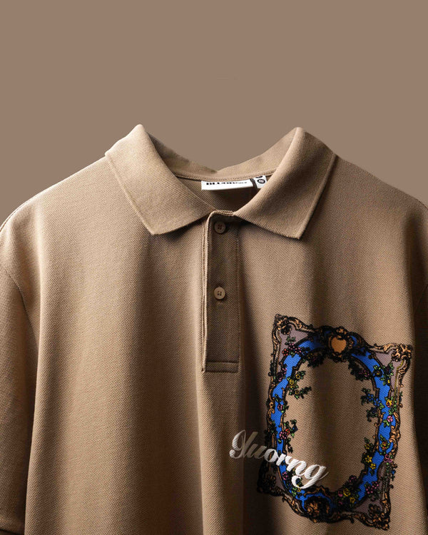 Close-up of the back of a beige polo t-shirt featuring a large ornate baroque-style graphic with a blue arch, gold scrollwork, and floral details.