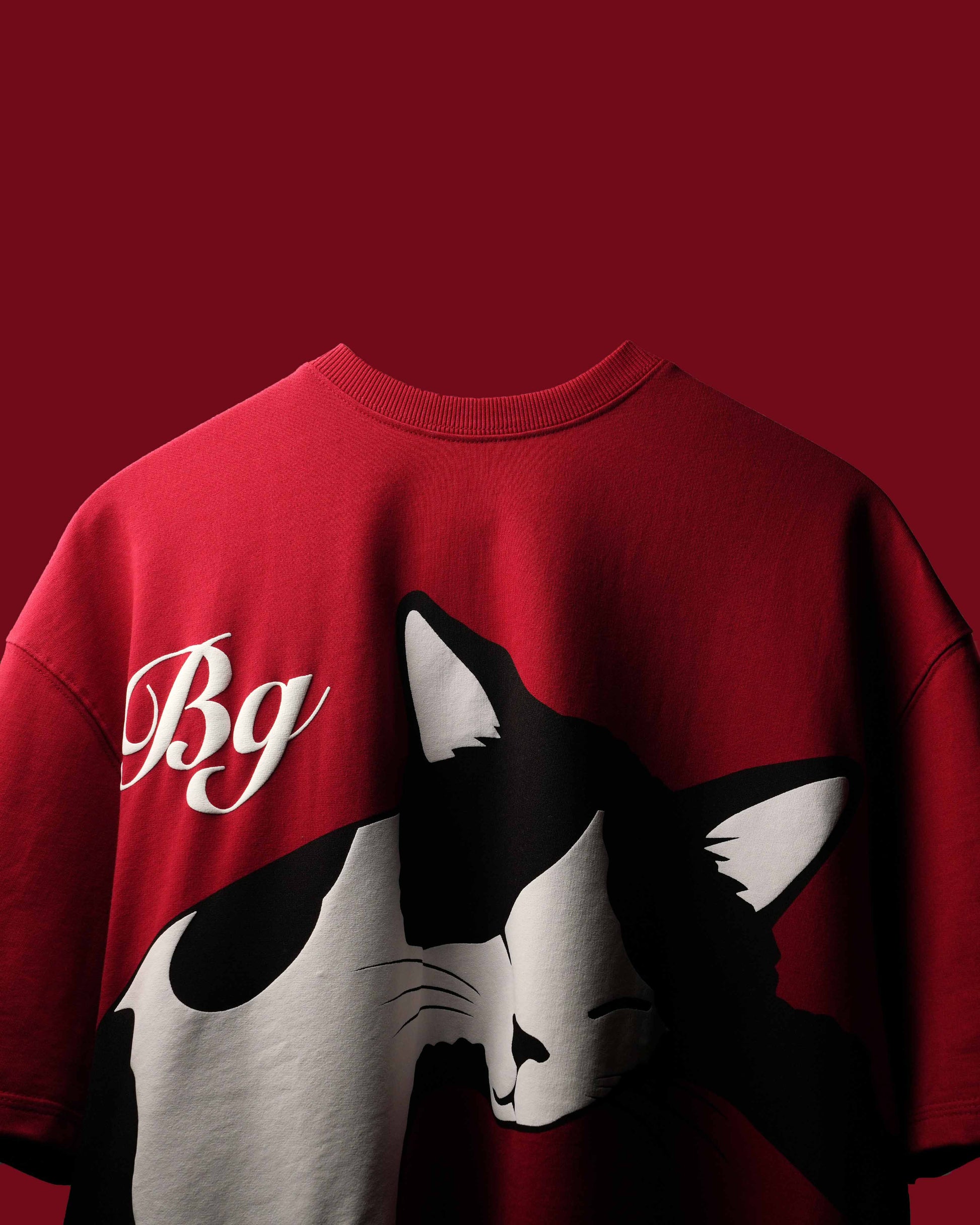 A red crewneck sweatshirt featuring a large graphic print of a black and white cat on the front, alongside a white cursive 'Bg' logo on the left chest, set against a solid dark red background.