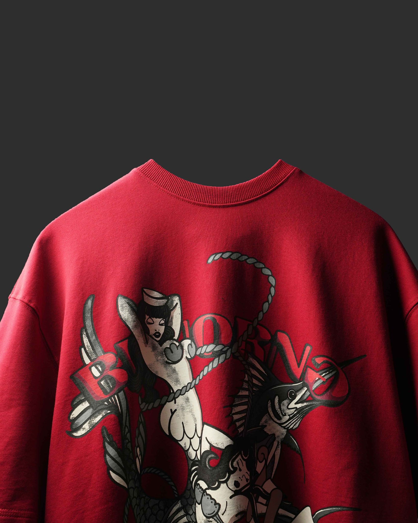 A close-up of a red crewneck sweatshirt featuring a large graphic print on the front with a tattoo-style mermaid, fish, and the word 'BORN'.