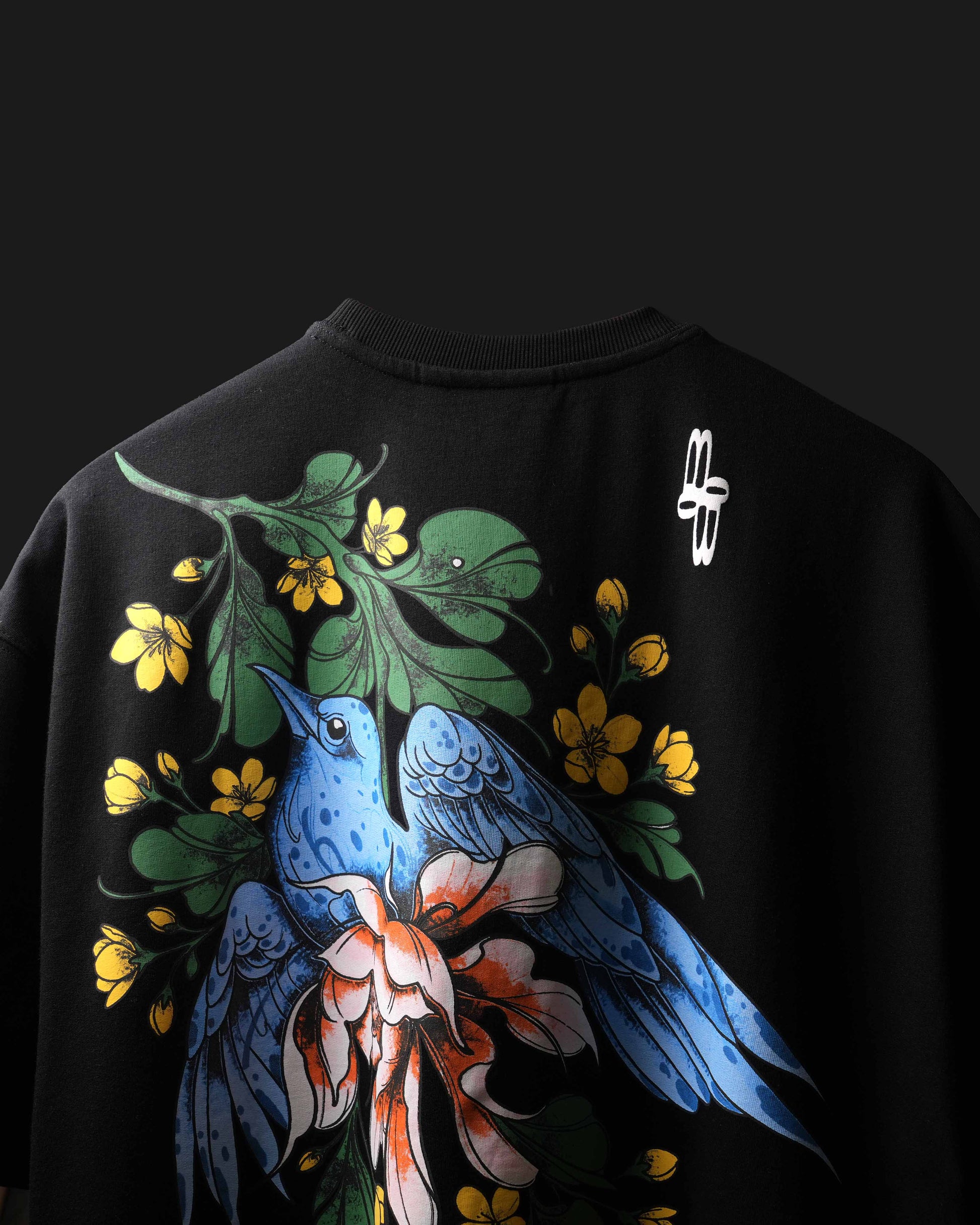 A black crewneck t-shirt featuring a large graphic print of two blue birds, green leaves, yellow flowers, and a pinkish-orange flower on the front. A small white logo is printed on the upper right shoulder area.