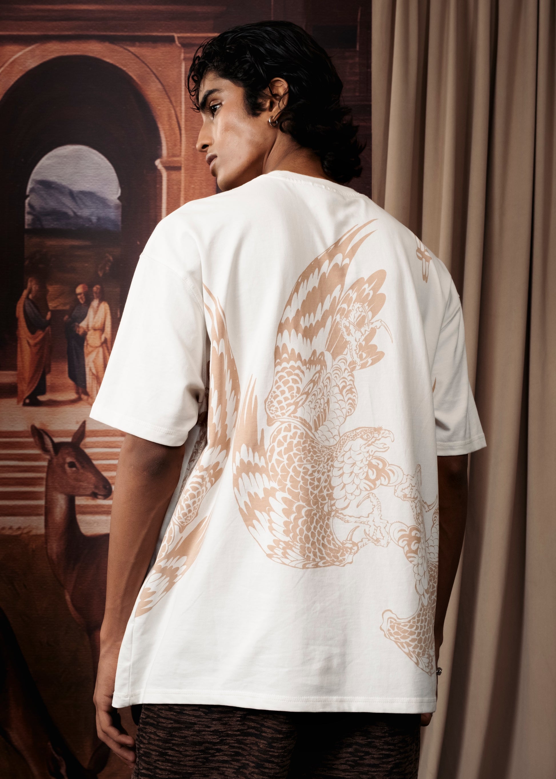 A man wearing a white short-sleeve t-shirt with a large beige phoenix graphic on the back, shown from behind in an indoor setting with a painting and curtain in the background.