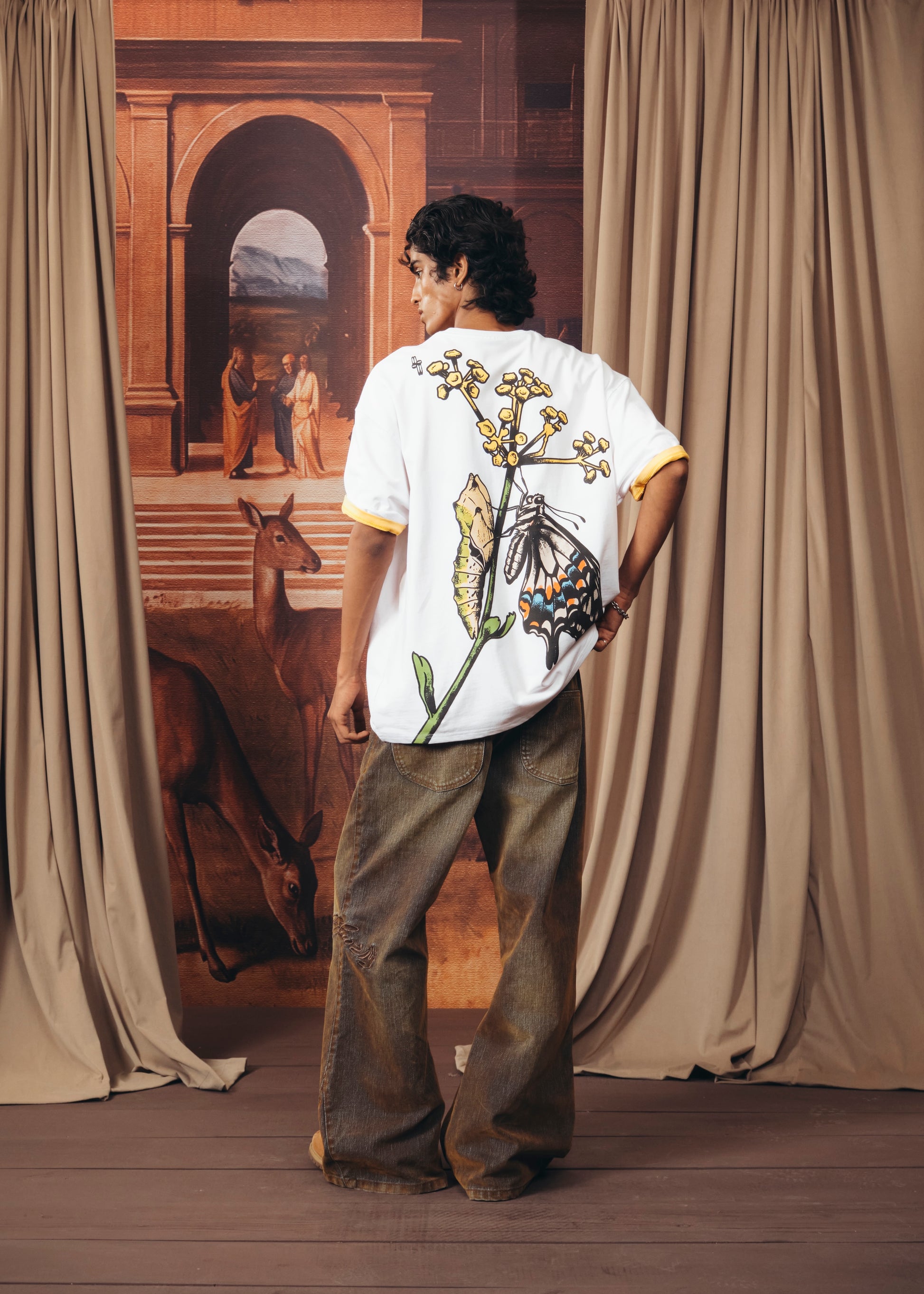 A model stands with their back to the camera wearing a white t-shirt with a large butterfly and floral graphic on the back, paired with brown wide-leg jeans. The setting features beige curtains framing a painted backdrop of an archway and a horse.