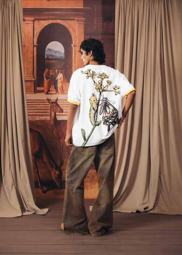 A model stands with their back to the camera wearing a white t-shirt with a large butterfly and floral graphic on the back, paired with brown wide-leg jeans. The setting features beige curtains framing a painted backdrop of an archway and a horse.