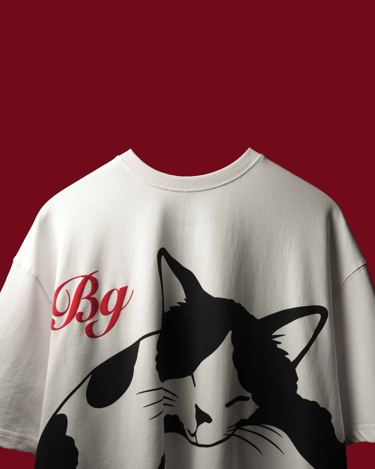 A white oversized crewneck t-shirt featuring a black cat graphic and red cursive 'Bg' lettering on the chest, set against a solid dark red background.