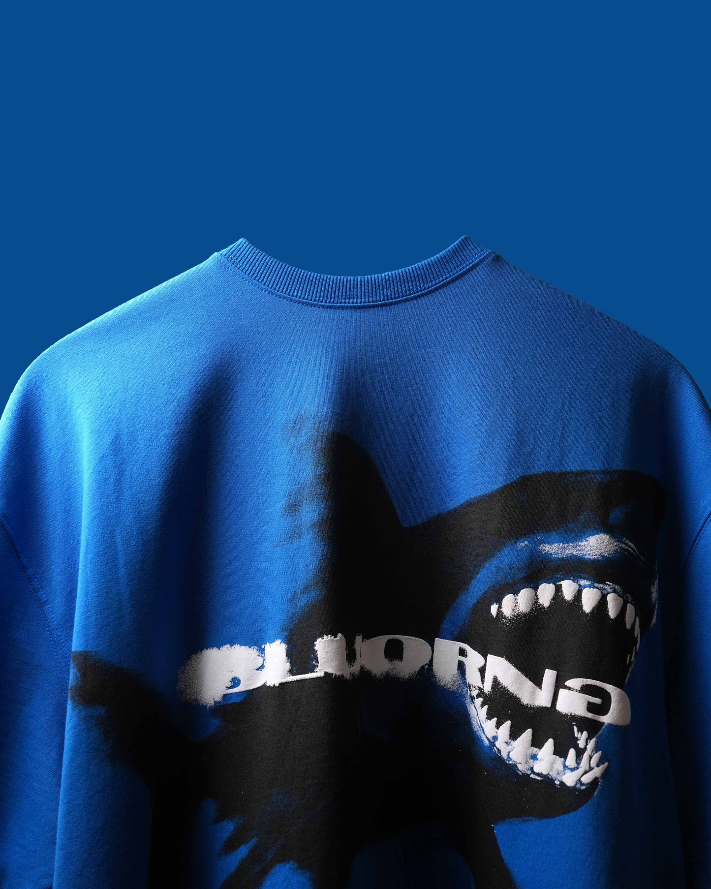 A close-up of a bright blue crewneck T-shirt featuring a black shark graphic with white teeth and the word 'BLUORNG' in white across the chest, set against a solid blue background.