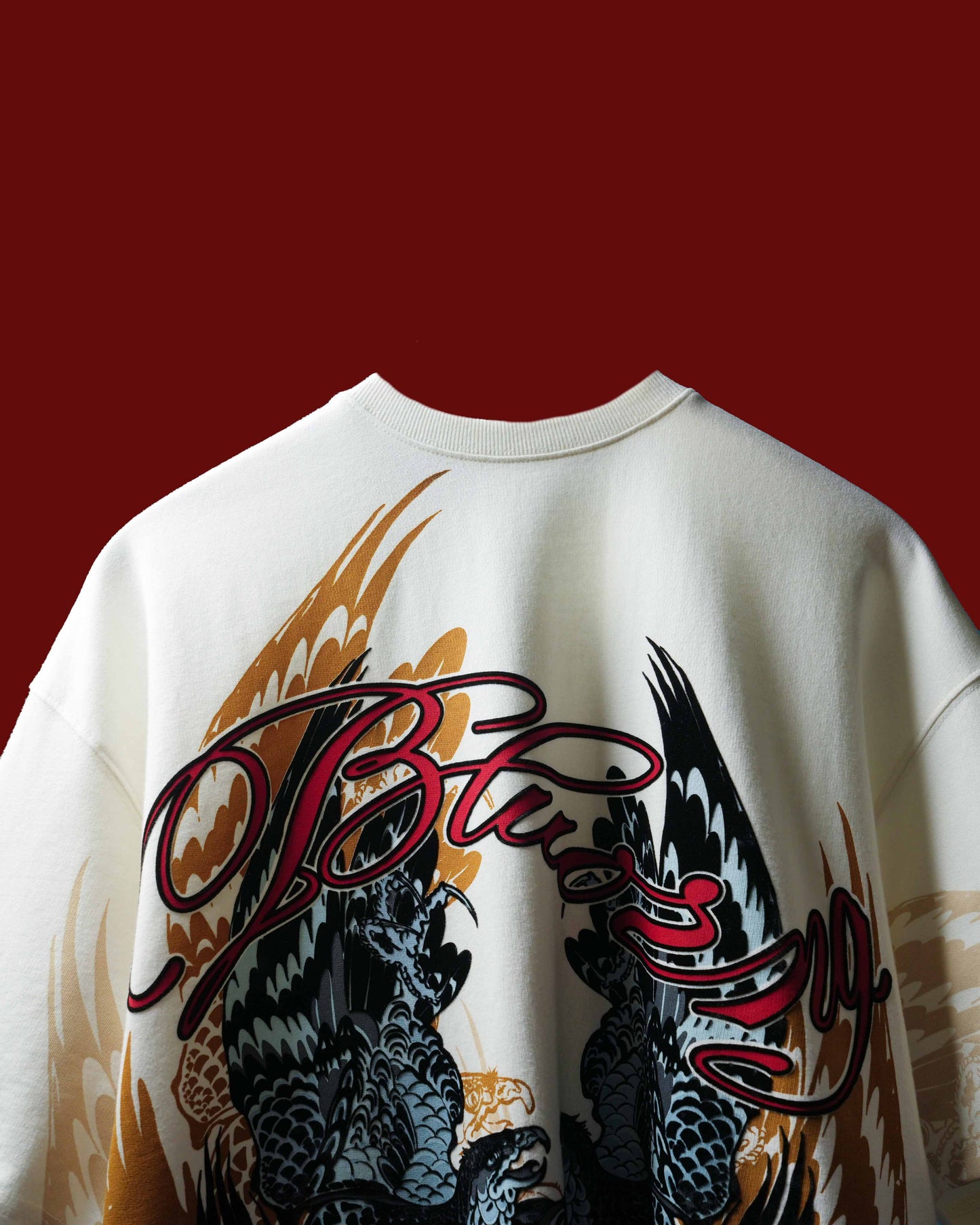 An off-white crewneck sweatshirt featuring a large graphic print of two black and gray eagle wings with gold flame accents and a red script logo across the chest, set against a solid dark red background.