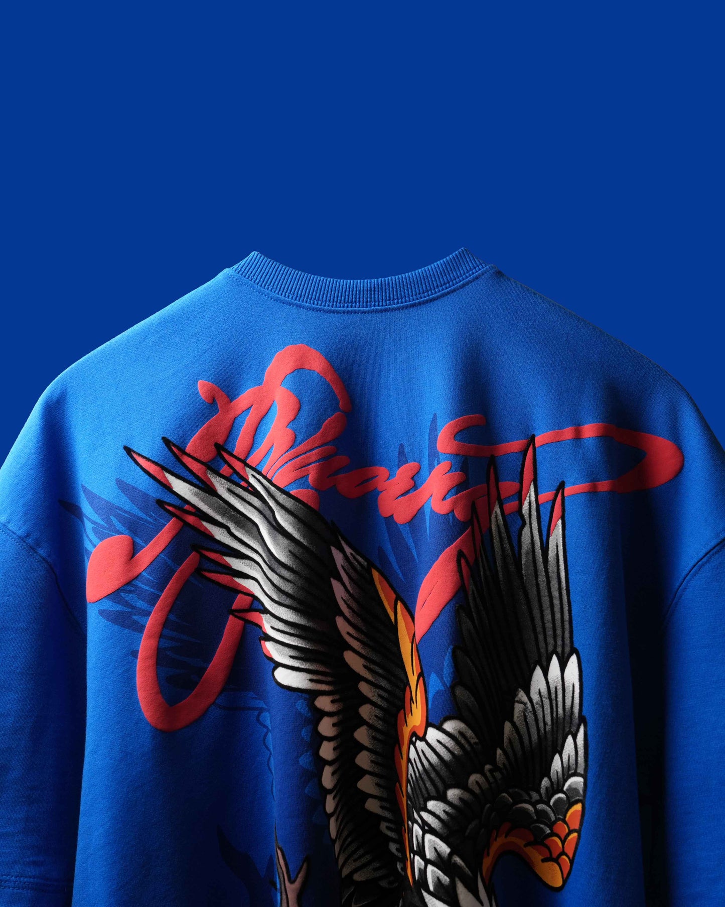 A close-up of the back of a blue crewneck sweatshirt featuring a large graphic print of a winged eagle and red stylized script lettering, set against a solid blue background.
